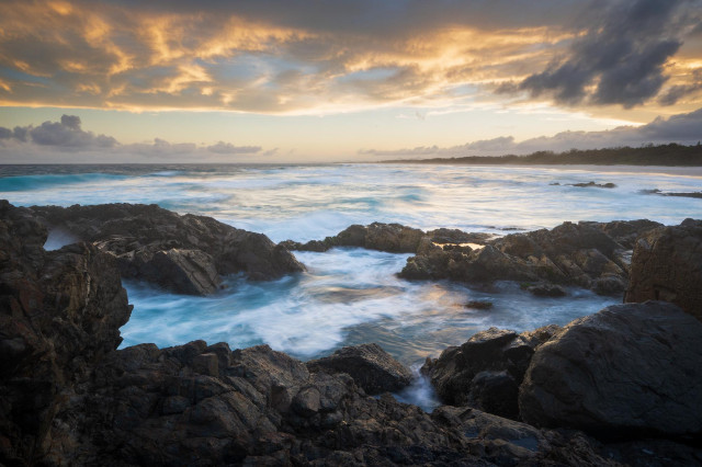 seascape with some rocks in the foreground encircling portion of the sea, then there is an open sea further back. In distance, there's a sliver of beach and hills and a sunset sky at the top reflecting in the water.