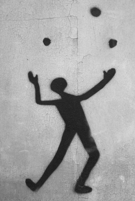 Auf einer hellgrauen Wand deren Putz/Farbe spröde abzublättern beginnt eine schwarze Figur, die dynamisch mit drei schwarzen, fast runden  Flecken jongliert. Es wirkt wie mithilfe einer Schablone auf die Wand gesprüht. 