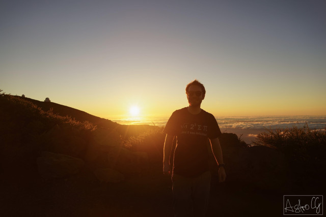 Me on top of a mountain above the clouds, just before sunset. The sun is setting in the back. On the horizon, big permanent telescopes can be seen.