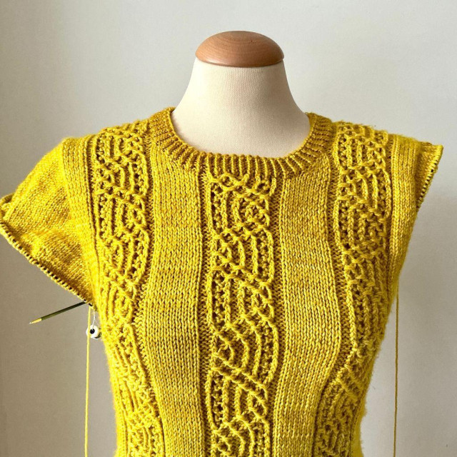 A sweater knitting project and a ball of bright yellow yarn, shown from the front on a mannequin against a white background. On the left a cap sleeve has been worked with short rows, the stitches are on a circular knitting needle. On the right, the short rows have just been started.