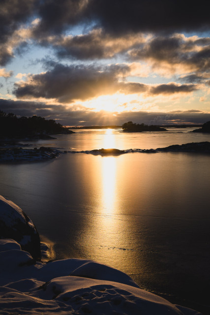 A vertically oriented photograph of the sun setting over the sea, which is covered by coarse ice. There are several small rocky islands and islet in the sea, some of which have trees on them and some are barren.

In the foreground some snow and bedrock from the near shore are visible. The upper part of the photo is a cloudy sky with blues and yellows of the late afternoon sky visible between the clouds. The sun shines through a gap in the clouds, creating a bright yellow line of reflection in the sea ice.
