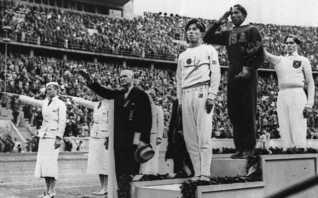 Officials and a German athelete give the Hitler salute as the sole African American gives a regular military salute and the Japanese athletes stands with his hands at his side at a medal ceremony during the 1936 Summer Olympics.