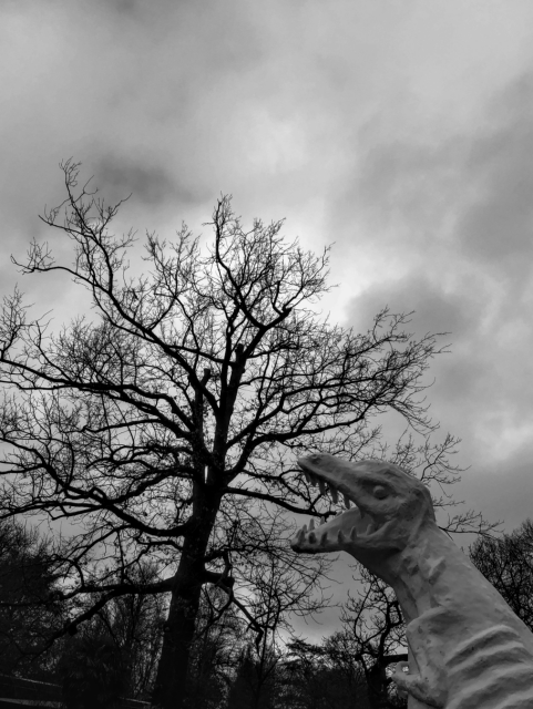 Black and white photo. Bare tree silhouetted against a clody sky. In the lower right of the frame is a white fierce dinosaur statue, mouth open and with sharp teeth. It appears in the image like the dinosaur is about to bite into the tree.