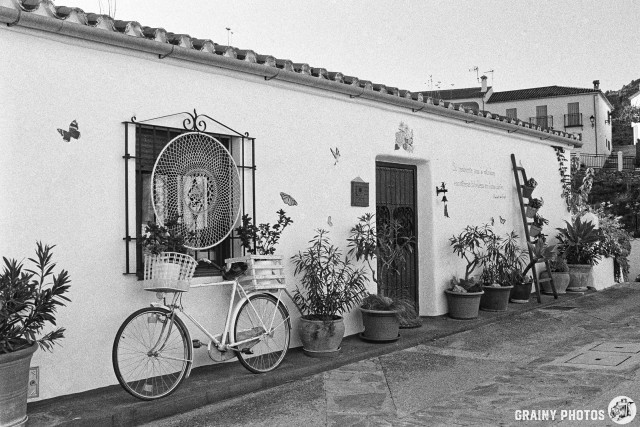 A charming white-washed exterior features a decorative window with a dreamcatcher, surrounded by lush potted plants and butterflies. A vintage bicycle leans against the wall, complemented by an inviting, rustic atmosphere.