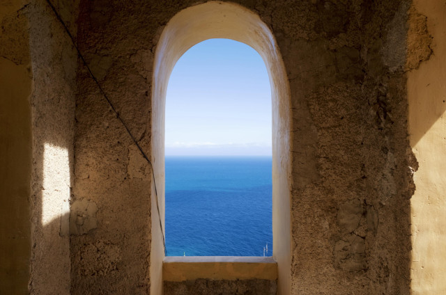 A view through an arched window showcasing a bright blue ocean and clear sky. The interior walls are textured and weathered, with a shadow cast on one side.