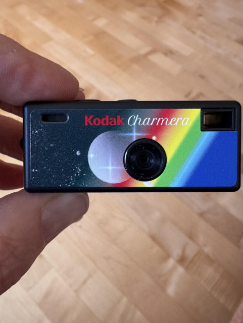 A hand is holding a Kodak Charmera camera, featuring a colorful design with a rainbow and star motifs. The camera is positioned against a wooden surface.