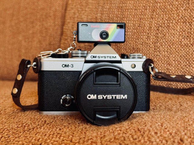 A new OM-3 camera with a lens cap on, featuring a Kodak Charmera camera mounted on top. The OM-3 camera is resting on a sofa with a textured brown surface, and has a brown leather strap attached.
