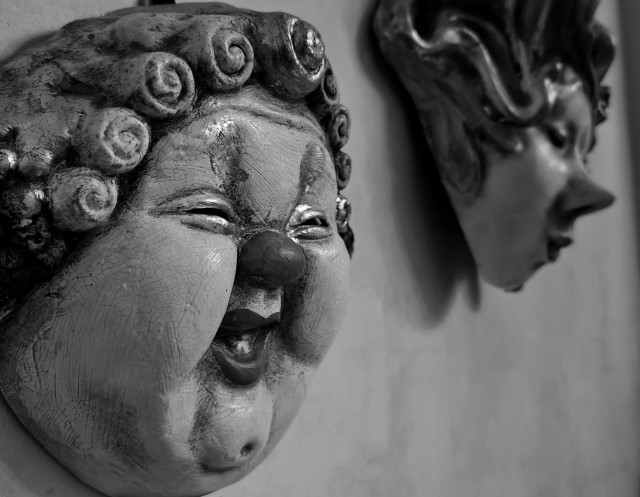 A black and white photo of two carnival masks, one in front with a tiny round nose and curly hair, grinning and another blurred in the background in profile.