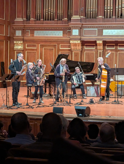 An Anthony Braxton led unit plays an experimental composition in a wooden theatre.  Three saxophones, one grand piano, one upright bass, and one accordion.