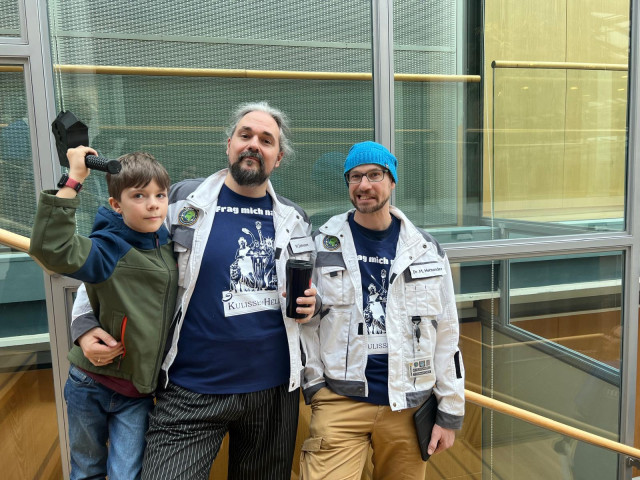 Der Co-Founder, flankiert von einem etwa zehnjährigen Jungen und einem jungen Mann mit Brille, vor einer Glaswand. 