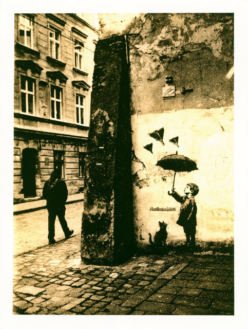 A lith print made in the center of Lviv. The composition is divided by the rough corner of an old building: on the left, a blurred passerby moves along a cobblestone street framed by historic façades; on the right, a cracked and peeling wall carries a powerful stencil.

The graffiti depicts a small boy holding an open umbrella above his head, protecting himself — and a cat at his feet — from attacking combat drones, the kind that strike Ukraine daily. The umbrella becomes a fragile yet symbolic shield against a very real and ongoing threat.

The lith process intensifies the scene with coarse grain, deep shadows, and warm sepia tones