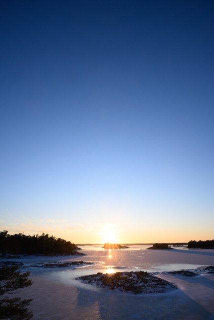 A vertically oriented landscape photography of the sun setting over a frozen over archipelago. The sky is taking up the vast majority of the vertical space in the photo, showing a gradient from yellow/orange at the horizon, to light blue, to deep darker blue at the top edge. The sky is entirely clear, apart from some very small, sparse clouds near the horizon.

The archipelago consists of a frozen over sea with just a little bit of snow on the ice, and a number of small rocky islands. The islands toward the horizon are predominantly covered by trees and the ones nearer the foreground are bare rock.

The sun is in the center of the frame, just above the horizon, casting a pale golden light over the landscape and creating deep shadows where it shines on the islands. There are golden reflections in the ice from the setting sun.