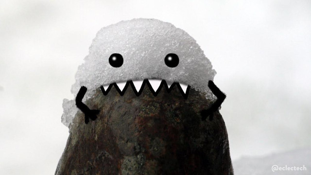 A close up photo of a small stone with a dollop of snow on the top. The background behind is blurred white snow. The snow dollop has arms holding on to the stone, cartoon eyes, and pointy teeth as if it is chomping on the stone. 