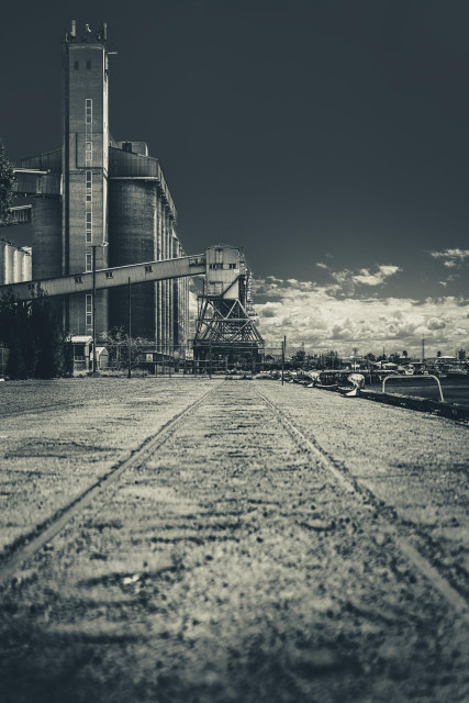 Monochrome image of a deserted industrial site with train tracks leading toward large silos and machinery. Cloudy skies create a sombre, nostalgic atmosphere.