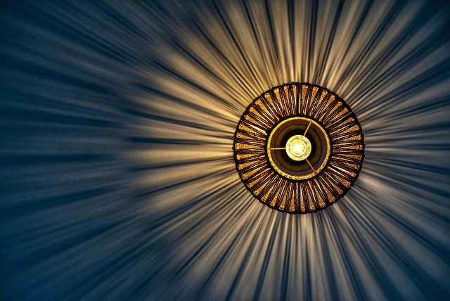 Das Bild zeigt eine kreisförmige Deckenleuchte in einer Nahaufnahme von direkt unten, die ein spektakuläres Licht- und Schattenspiel an die Wand wirft.
Im Zentrum der Komposition befindet sich die leuchtende Glühbirne, die ein warmes, gelbliches Licht ausstrahlt. Sie ist von einem runden Lampenschirm umgeben, der aus vielen dünnen, dunkelbraunen oder schwarzen Stäben besteht, die strahlenförmig von der Mitte nach außen verlaufen. Diese Stäbe sind durch zwei konzentrische Ringe miteinander verbunden, was dem Schirm eine strukturierte, fächerartige Optik verleiht.
Das markanteste Merkmal ist der dramatische visuelle Effekt: Das Licht der zentralen Quelle dringt durch die Zwischenräume der Stäbe und erzeugt hunderte von feinen, hellen Lichtstrahlen, die sich symmetrisch und weiträumig über die gesamte dunkelblaue Wandfläche ausbreiten. Zwischen diesen Lichtstreifen entstehen ebenso feine, dunkle Schattenlinien, wodurch ein dynamisches Muster entsteht, das an eine strahlende Sonne oder eine Explosionszeichnung erinnert. Die tiefblaue Farbe der Wand bildet einen starken Kontrast zum warmen, goldenen Schein des Lichts und verstärkt die Tiefenwirkung des Musters.