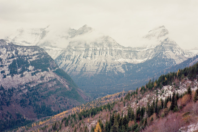 A few layers of mountains getting hazier into the distance. The near mountain slope has the reds, greens, and yellows of Fall. The middle ridge is quite steep has more exposed rocks with a dusting of snow. The farther mountain can be seen all the way from the dense trees of the valley to the peaks playing hide and seek with clouds and cover in snow with jagged rock and ridges emerging from the clouds and snow. Shot with a Voightlander BessaR2 and 7Artisan35f2 lens, on KodakEktar100, and developed with KodakC41 by Shom Bandopadhaya. Licensed under Creative Commons Attribution-NonCommercial-ShareAlike (CC BY-NC-SA).
