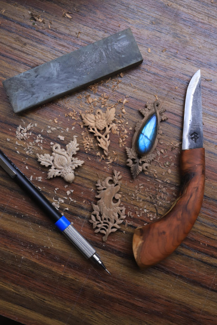 Atelier de Lucie BRAILLON de l'atelier de La lettre aux ours situé en Normandie. Quatre sculptures miniatures en bois (pendentifs) en cours de travail sur son établi, entourées de copeaux et d'outils (un criterium, une pierre à affuter et un couteau de La forge d'Asgeir). Les sculptures reprennent des thématiques diverses autour de la vie sauvage et de la nature : un loup, des arbres, des plantes, des ronces, un gland et des feuilles de chêne...