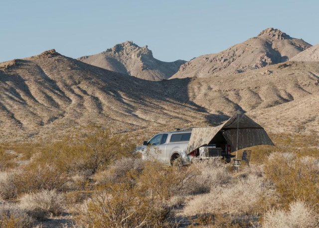 A color landscape photo showing a gray pickup truck with a homemade awning attached to the back and a chair under the awning set among yellow-green creosote bushes. In the background are tall, pretty much barren hills that rise to three distinct peaks under a clear blue sky.