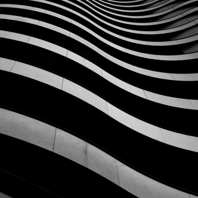 Black and white photo looking up at a section of a building's face, curved balconies swell out from a pin-striped wall, little visible but dully reflective waved stripes between black shadowed undersides.