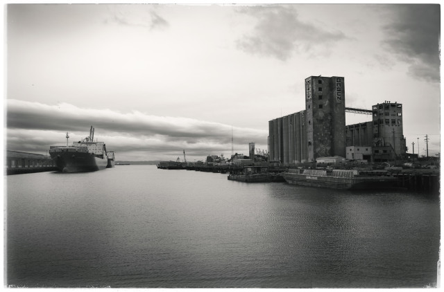 A moody, monochrome picture of the San Francisco Bay waterfront, taken yesterday, for 4:11 p.m., a few hours before the start of a promised two weeks worth of rain.

Above, stormy skies.

On the left, Pier 80 with two ships docked alongside the pier's single story shed.

On the right, Pier 90, with a low slung barge docked next to seven story grain silos. This pier has been crumbling for decades and it days are numbered. 

In the center, the broad Islais Inlet. Here, the waters of a creek that begins 3-4 miles away, widen and flow out to the bay.