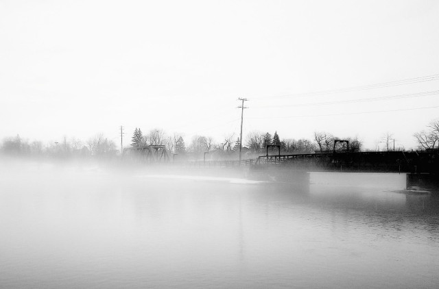A train bridge is seen partially obscured by fog crossing a river. This is a high contrast black and white image.