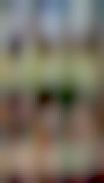 A photo of a dense tuft of snowdrops growing on a carpet of dead leaves and twigs. Mostly bare trees are visible indistinctly in the background, as well as a bit of blue sky peeking through the clouds.