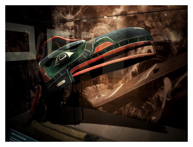 A carved wooden mask painted in red and green, in the elongated shape of a bird-like head and open beak, on display in a glass case. Filling the wall in the background is a sepia-toned photograph of many people wearing ceremonial masks of a similar kind.