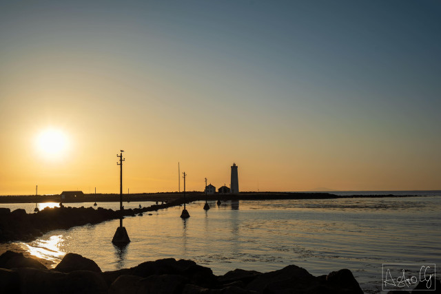 Sunset over calm sea with a lighthouse and several masts in the water