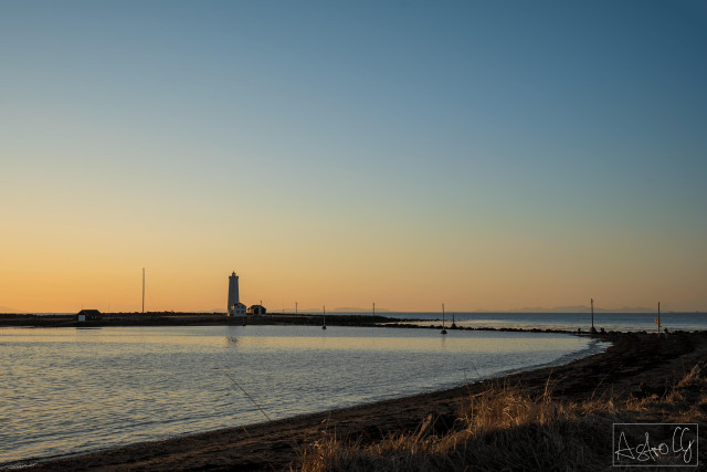 Coastal landscape with calm water, lighthouse, and buildings on the horizon at sunset
