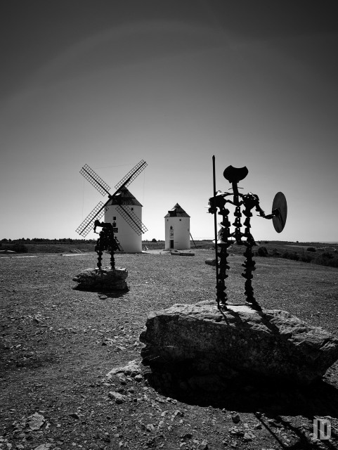 La imagen en blanco y negro muestra un paisaje rural con varios molinos de viento tradicionales. En primer plano, se ven dos esculturas metálicas que representan a Don Quijote y Sancho Panza, situadas sobre rocas. Al fondo, destacan los molinos bajo un cielo despejado, creando un contraste marcado entre las siluetas oscuras de las figuras y la luminosidad del horizonte.