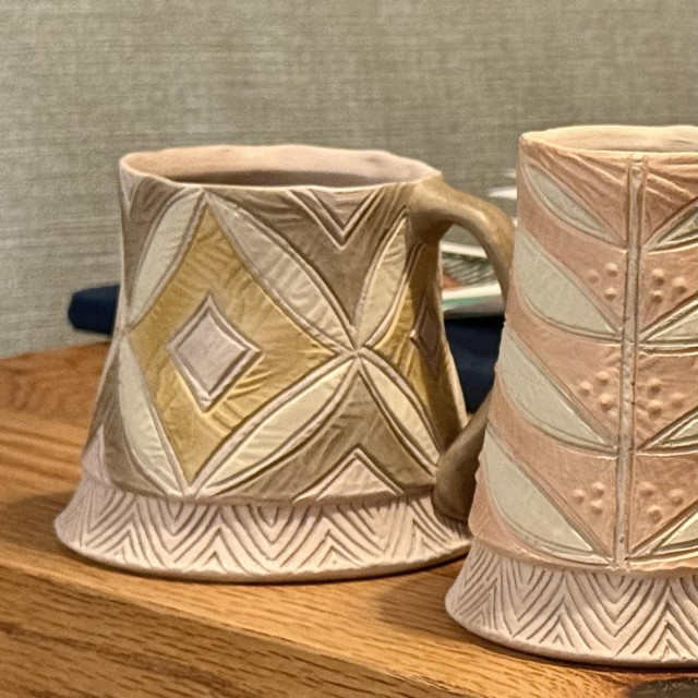 Two unfired ceramic mugs with intricate geometric patterns and textures, one featuring gold and gray colors, and the other in soft pink tones. They are placed on a wooden table with a blurred background.