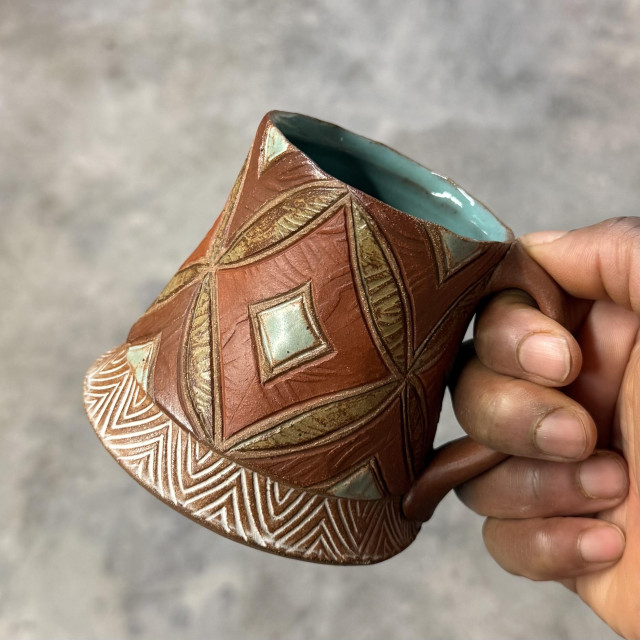 My hand holding a finished ceramic mug with carved diamond patterns painted in earthy tones of brown and turquoise. Blurred in the background, the gray cement floor in my studio. 