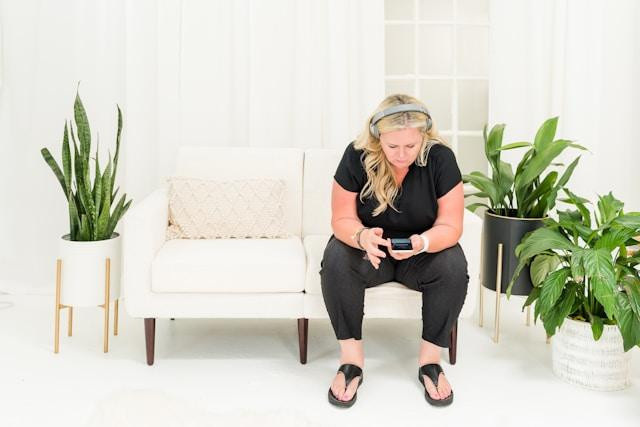 Bright photo. A person sits on an off-white couch in a pale room with some plants. Their feet are on the floor. They sit with elbows on the knees, looking down at a phone with over-the-ear headphones. Long blond hair and pink skin wearing all black. Thong sandals.