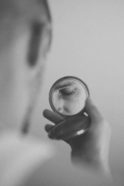 A vertical, grainy black-and-white photograph with a shallow depth of field.

In the foreground, a hand holds a small, open circular compact mirror. The reflection in the mirror shows a sharp, detailed close-up of a person's closed eye and eyebrow. The rest of the image is softly blurred, including the person's hand in the bottom right and the back of a head and ear in the upper left background. The overall mood is intimate and contemplative.