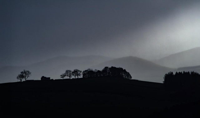 Black and white landscape looking over to some blurry stormy hills, the mid and fore grounds are a black silhouette of some  trees and a castellated building.