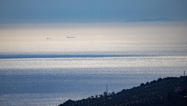 A hillside slanting down just below centre right to middle bottom. On the sunlit  Mediterranean sea are two ships and behind that coming out of the haze towards the right are the Riff mountains in Morocco, Africa.