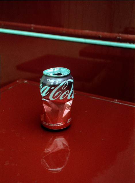 The image shows a crushed Cherry Coca-Cola can placed upright on a glossy red metal surface. The can is partially crumpled in the middle, its aluminum body sharply folded inward, yet the top remains open and intact. The deep red of the can blends almost seamlessly with the equally saturated red background, creating a monochromatic scene interrupted only by the white Coca-Cola lettering and the pale turquoise tone of the can’s rim.

The reflective surface beneath mirrors the distorted shape of the can, adding a soft, warped reflection that echoes its crushed form. In the background, slightly out of focus, a horizontal metal bar runs across the frame, reinforcing the industrial, minimal atmosphere.