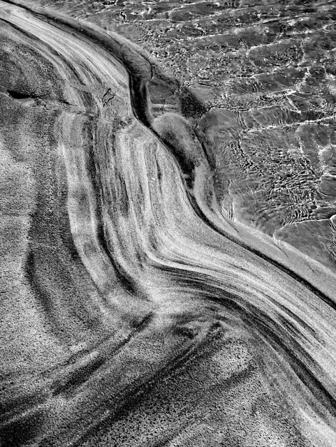 Hochformatige schwarz-weiße Aufnahme am Strand von Seilebost, Hebrides, Scotland. Der Strand an der Wasserkante ist vom Wasser geformt, in der rechten oberen Hälfte ist Wasser in Bewegung, der Rest des Bildes ist Sand, verschieden Bögen und Linien in unterschiedlichen Farbtönen bilden eine interessante Struktur.

Portrait-format black-and-white photograph taken on Seilebost Beach, Hebrides, Scotland. The beach at the water's edge has been shaped by the water; in the upper right-hand half, the water is in motion, while the rest of the image is sand, with various arcs and lines in different shades forming an interesting structure.