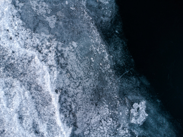 Is på en sjö sett uppifrån. Till vänster i bilden  är äldre is som är nästan helt vit och inte går att se igenom. Till höger i bilden är ny, tunnare is som är helt transparent och det enda som syns är det mörka vattnet. Övergången mellan de två isarna delar bilden diagonalt.