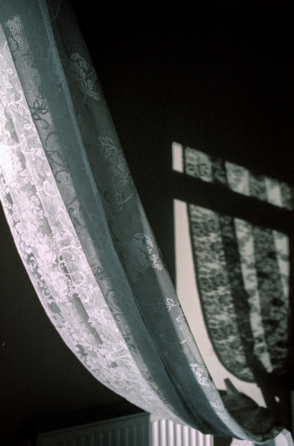 A softly lit interior scene with a lace curtain drifting gently across the frame. The sheer fabric, detailed with delicate floral patterns, catches the light, creating a luminous contrast against the darker room behind it. On the right side, the blurred shadow of a window frame falls across the wall, forming geometric shapes that echo the curtain’s curves. Below, a radiator is partially visible, grounding the composition in a quiet domestic space. The image feels intimate and contemplative, balancing texture and shadow with a subtle, muted color palette.