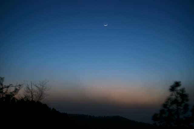 The evening twilight sky as a dust storm is easing up. The colors are subdued and the nearby trees are blurred in the exposure as they whip back and forth in the strong wind.  Venus is visible below the crescent moon, about 1/3 of the way up from the very bottom of the frame. Mercury is visible next to the crescent moon. 