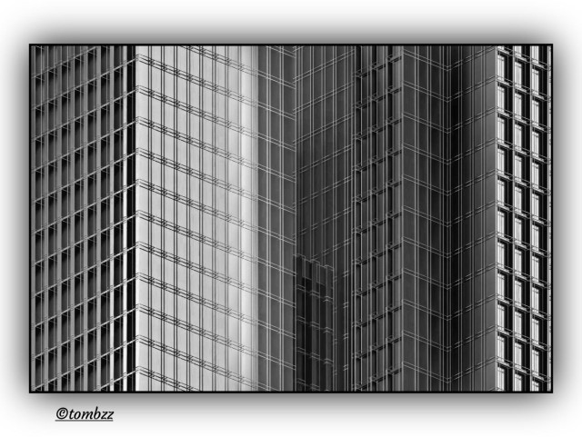 Black and white photo showing several tall buildings with modern, geometric architecture. The frame is tight and angled upward, so the facades fill the image with repeating rectangular windows and vertical structural lines. The buildings lean slightly toward the center, which adds depth and a sense of scale. Strong contrast highlights sharp edges and the difference between lit and shadowed surfaces. The whole scene turns into an almost abstract pattern built from rhythm, light and shadow.