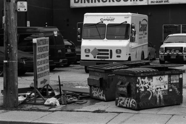 A small, chaotic industrial parking lot, with debris crowded by the sidewalk, two graffiti covered dumpsters, a placard boasting cheap mattresses and furniture, and a lopsided delivery truck. 