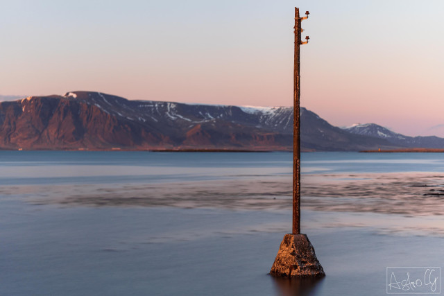 A single pole on a small platform stands in the water with a mountain range in the background during twilight