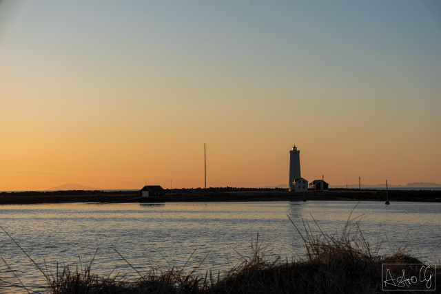 Lighthouse and buildings on the shore at sunset with calm water in the foreground