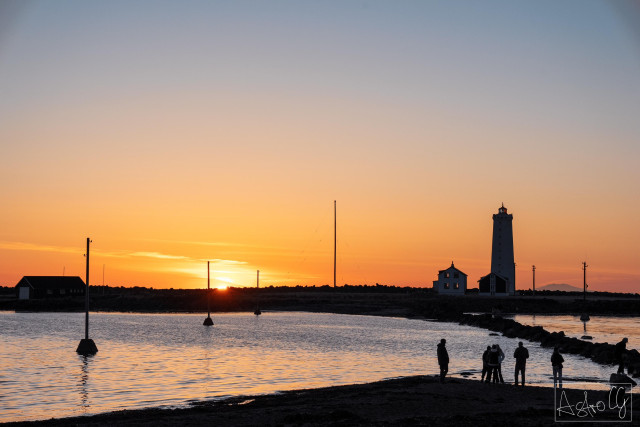 Silhouettes of people on the shore with a lighthouse and posts on the water at sunset