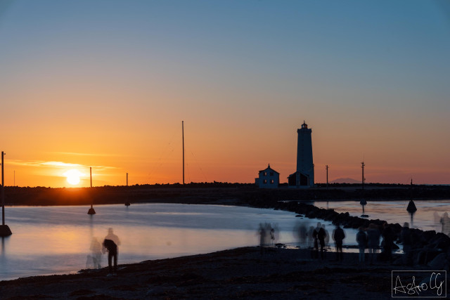 Blurry silhouettes of people on the shore with a lighthouse and posts on the water at sunset
