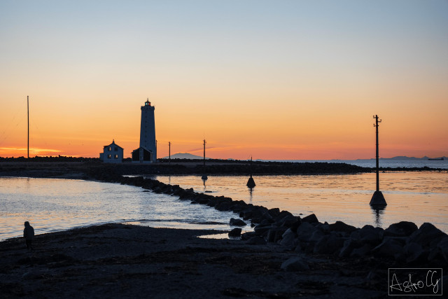 Lighthouse and buildings on the shore at sunset with calm water and rocks in the foreground