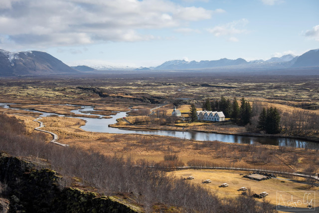 River landscape with meadows, trees, and a small white building in front of mountains under a cloudy sky