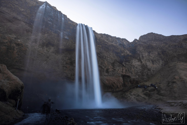 Waterfall plunging from a high cliff into a river with rocky banks under a blue sky
